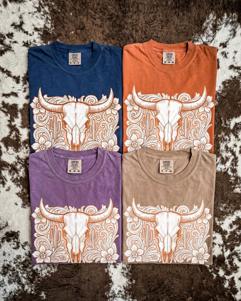 4 folded shirts on a brown and white cowhide rug. Rust, Navy, Purple, and Brown with a tooled skull design on them.