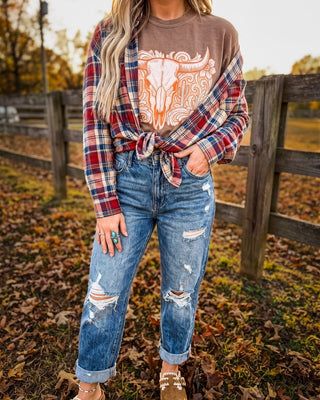 A woman wearing a brown graphic t-shirt with a red and blue plaid flannel shirt tied at the waist, and distressed blue jeans, standing outdoors in front of a wooden fence.