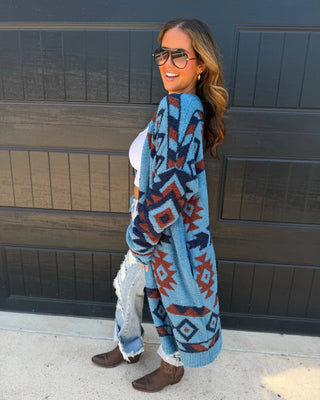 A woman wearing a blue knit cardigan with a red and dark gray geometric pattern, standing in front of a dark garage door.