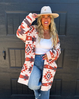 A woman stands in front of a black garage door wearing a cream and rust Southwestern patterned cardigan, a white tank top, blue jeans, a beige wide-brim hat, and gray boots.