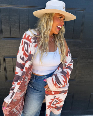 A woman wearing a white wide-brim hat, a white tank top, blue jeans, and a chunky knit cardigan with a red, brown, and cream Southwestern geometric pattern, standing in front of a black paneled door.