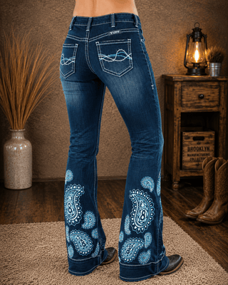 Rear view of a person wearing blue western flare jeans with white embroidered paisley patterns on the lower legs, standing in a rustic room with a wooden cabinet, boots, and a lamp.