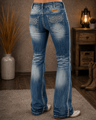 Rear view of a person wearing blue western-style flare jeans with decorative stitching and whiskering, standing indoors on a rug near a wooden cabinet and lantern.
