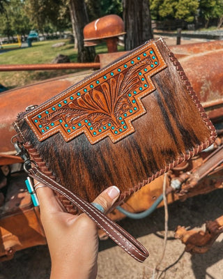 Brown leather clutch with intricate designs held by a hand, with a rustic background.