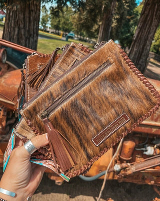 Brown leather clutch with fringe and brand logo held by a hand outdoors.