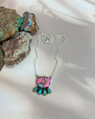 A silver pendant necklace featuring a pink and turquoise design, displayed on a draped white fabric next to decorative rocks.
