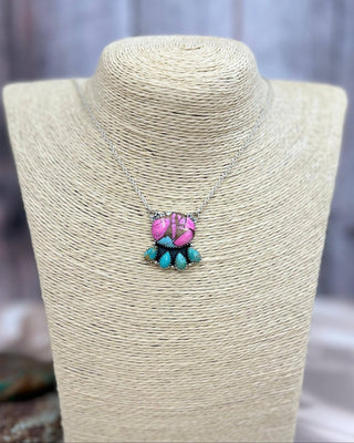 A silver chain necklace with a pendant, displayed on a beige textured jewelry bust. The pendant features a pink and purple patterned top with four turquoise blue dangling beads below.