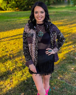 A woman posing outdoors in a grassy field at sunset, wearing a bohemian-style outfit consisting of a leopard print and sequined jacket, a black mesh top, a black fringe mini skirt, layered necklaces, a large green ring, and bright pink cowboy boots.
