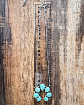 Turquoise colored pendant necklace with silver chain shown next to ruler for scale on wood surface.